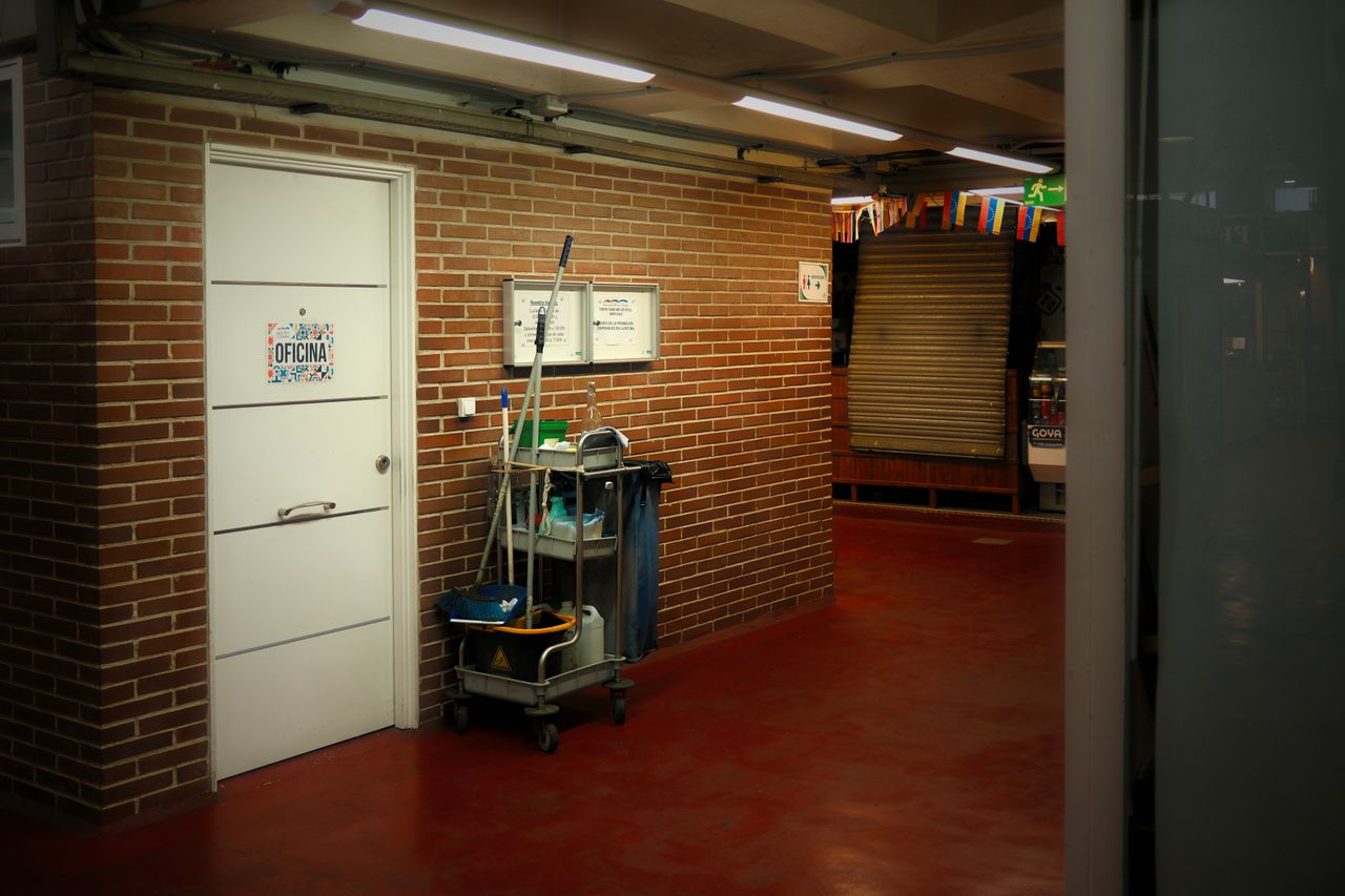 Un couloir industriel bien éclairé, avec un chariot de nettoyage et des fournitures près de la porte d’un bureau.