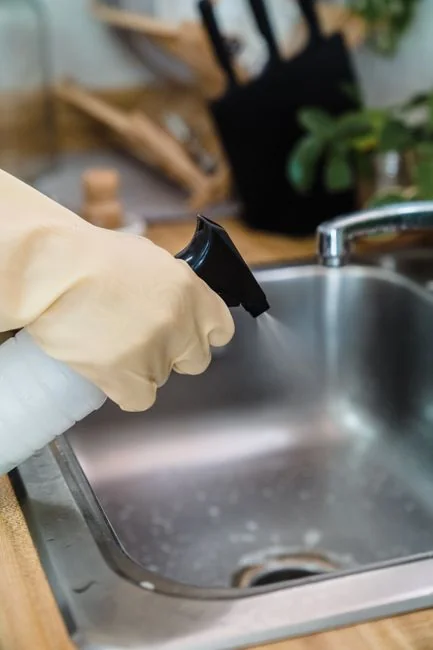 A person wipes a laptop screen using a blue cloth and spray bottle on a wooden table for hygiene.